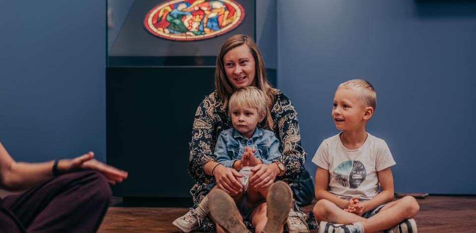 Een moeder en twee kinderen zitten in een museumzaal.