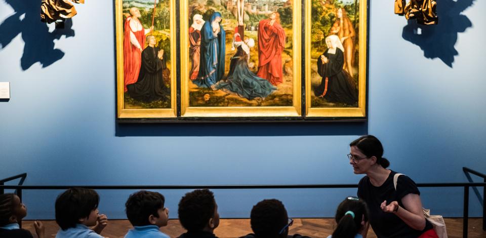 Een groep kinderen zit op de grond en kijkt naar het drieluik 'Calvarie met stichterspaar' van Quinten Massijs.